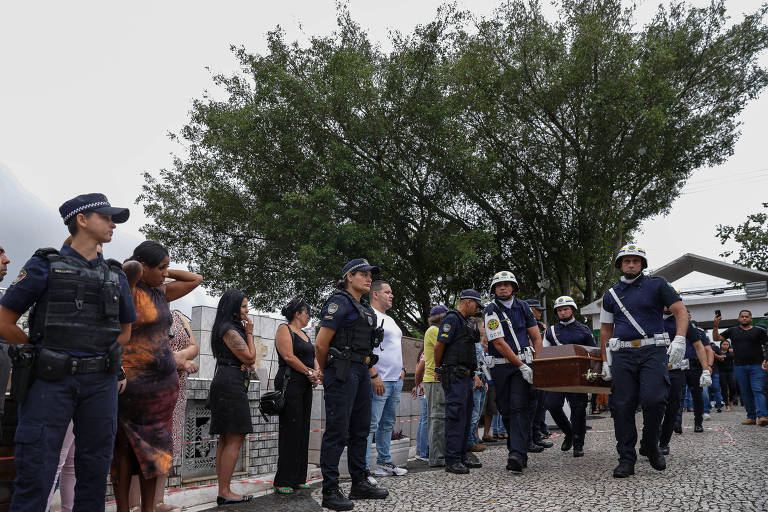 A imagem mostra uma cerimônia fúnebre ao ar livre， com várias pessoas em pé ao longo de um caminho. Um grupo de homens， vestidos com uniformes de polícia， carrega um caixão. À esquerda， algumas pessoas estão visivelmente emocionadas， enquanto outras observam a cena. Ao fundo， há uma árvore grande e um edifício. O céu está nublado.