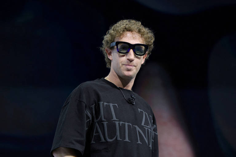 Um homem com cabelo cacheado e óculos escuros está em um palco, usando uma camiseta preta com texto. Ele parece estar se preparando para falar em um evento, com um fundo desfocado que sugere um ambiente de apresentação.