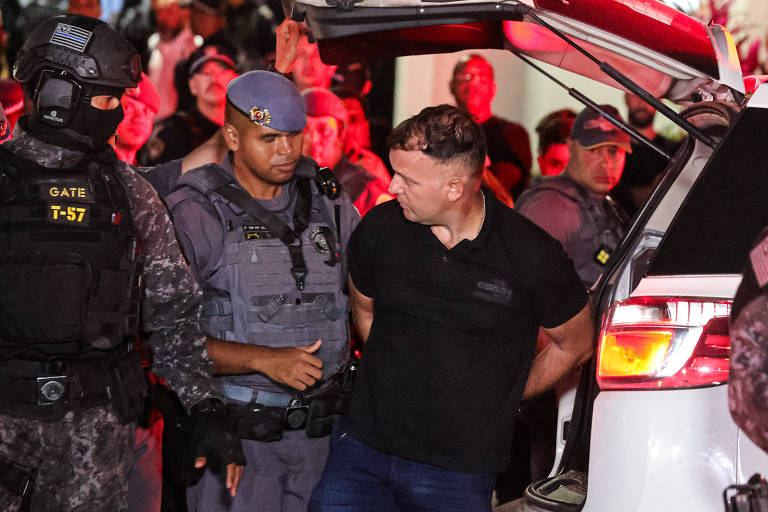 A imagem mostra um homem sendo escoltado por policiais em um ambiente noturno， com luzes vermelhas ao fundo. O homem， que está com as mãos algemadas， usa uma camiseta preta e parece estar sendo levado para um veículo. Dois policiais estão próximos a ele， um deles com um uniforme tático e máscara， enquanto o outro usa um boné e um uniforme da polícia. Há várias outras pessoas e policiais visíveis ao fundo.