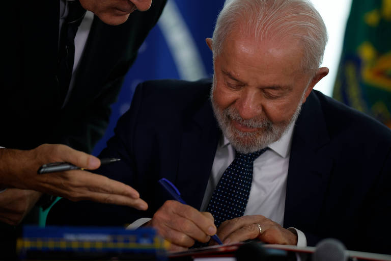 Lula， de cabelos grisalhos e barba， vestido com um terno escuro e gravata， está assinando um documento em uma mesa. Outro homem， com óculos e vestido de forma semelhante， está inclinado sobre ele， aparentemente discutindo ou orientando. Ao fundo， há bandeiras visíveis， incluindo a bandeira do Brasil.