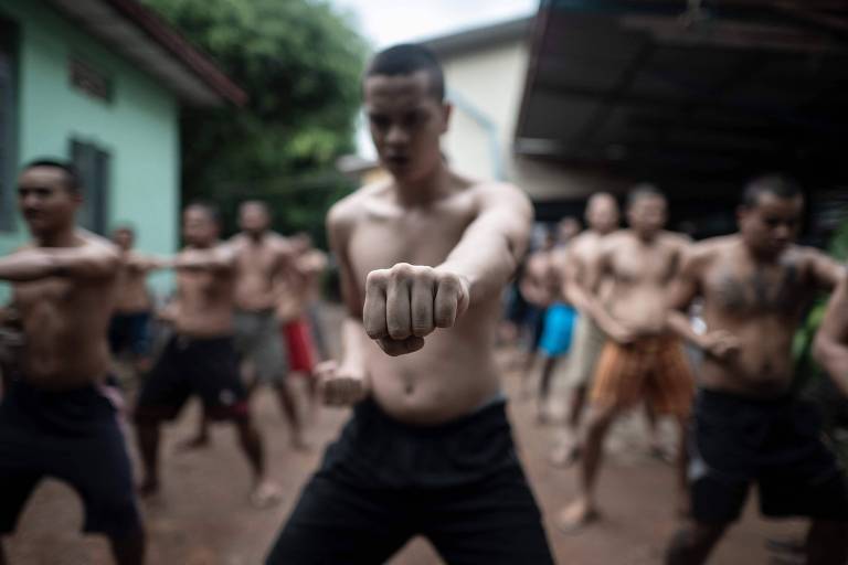 Um grupo de homens sem camisa está praticando artes marciais em um ambiente externo. O foco está em um jovem à frente， que está com o punho cerrado e em posição de ataque. Ao fundo， outros praticantes estão em posições semelhantes， todos aparentando concentração. O cenário é uma área com casas e vegetação ao redor.
