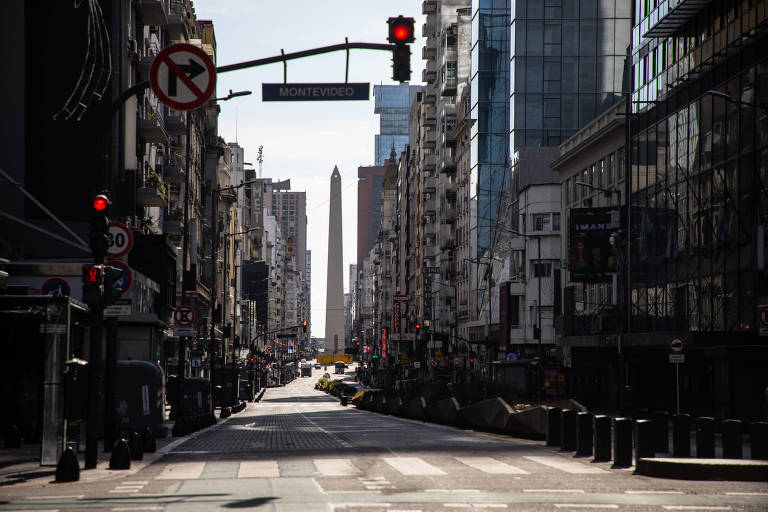 A imagem mostra uma vista da Avenida 9 de Julho em Buenos Aires， com o Obelisco ao fundo. A rua está vazia， com semáforos vermelhos e sinais de trânsito visíveis. Edifícios altos cercam a avenida， refletindo a arquitetura urbana da cidade. O céu está claro， indicando um dia ensolarado.