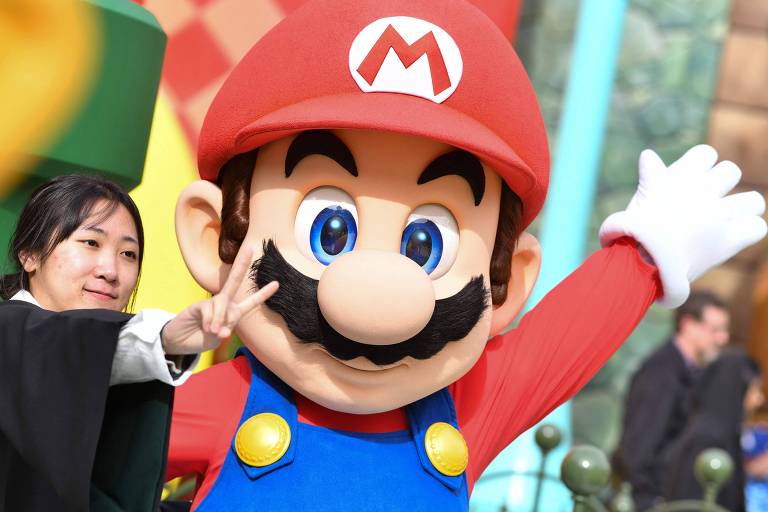 A imagem mostra uma pessoa vestida como o personagem Mario， da franquia de jogos， em um evento. O Mario está sorrindo e fazendo um gesto com a mão. Ao lado dele， há uma mulher que está posando ao lado do personagem， fazendo um número 2 com os dedos. O cenário é colorido e festivo.