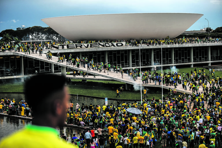 A imagem mostra uma grande multidão de pessoas reunidas em frente ao Congresso Nacional em Brasília. Muitas pessoas estão vestindo camisetas amarelas e verdes, segurando bandeiras do Brasil. Um grande banner com a palavra 'INTERVENÇÃO' está pendurado na estrutura do edifício. A cena é animada, com pessoas caminhando e algumas se reunindo em grupos. Ao fundo, há um lago e a arquitetura moderna do Congresso.