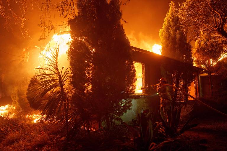 Bombeiro segura mangueira no ombro enquanto aponta para uma casa completamente tomada pelas chama. O fogo extrapola os limites do teto e atinge árvores ao redor