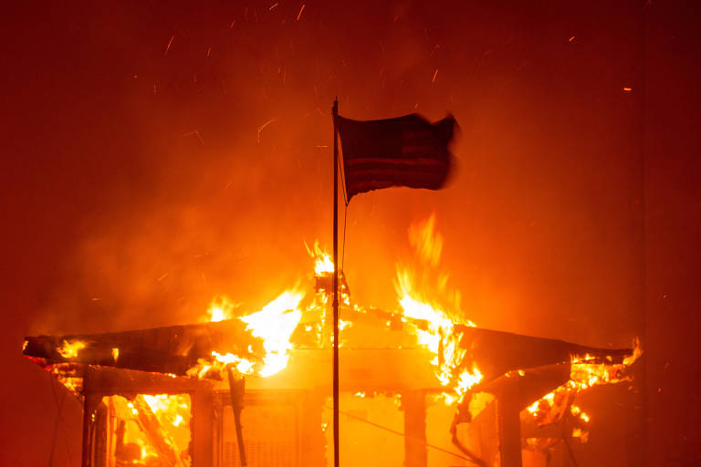 A imagem mostra uma estrutura em chamas， cercada por fogo e fumaça. Uma bandeira está visível， balançando ao vento， em frente à construção em chamas. O fundo é predominantemente laranja e vermelho devido ao fogo.