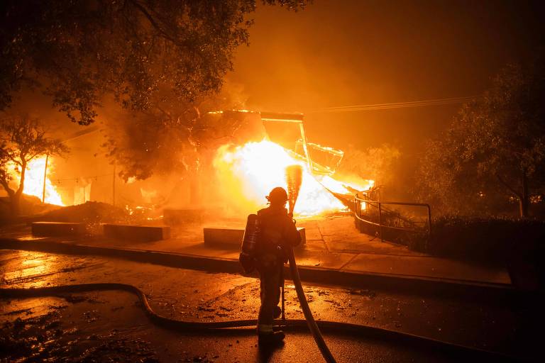 A imagem mostra um bombeiro em pé, segurando uma mangueira, diante de um grande incêndio que consome uma estrutura. As chamas são intensas e iluminam a cena, enquanto a fumaça densa se espalha pelo ar. O ambiente é escuro, com árvores visíveis ao fundo.