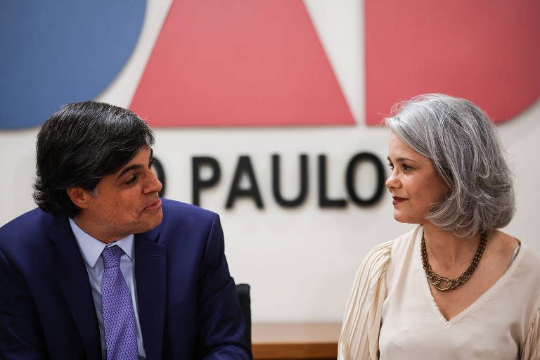 A imagem mostra duas pessoas sentadas em uma mesa, conversando. À esquerda, um homem com cabelo escuro e liso, vestindo um terno azul e uma gravata roxa. À direita, uma mulher com cabelo grisalho, usando uma blusa clara e um colar. Ao fundo, há um painel com as cores azul, vermelho e branco, e a palavra 'PAULO' em letras grandes e pretas.