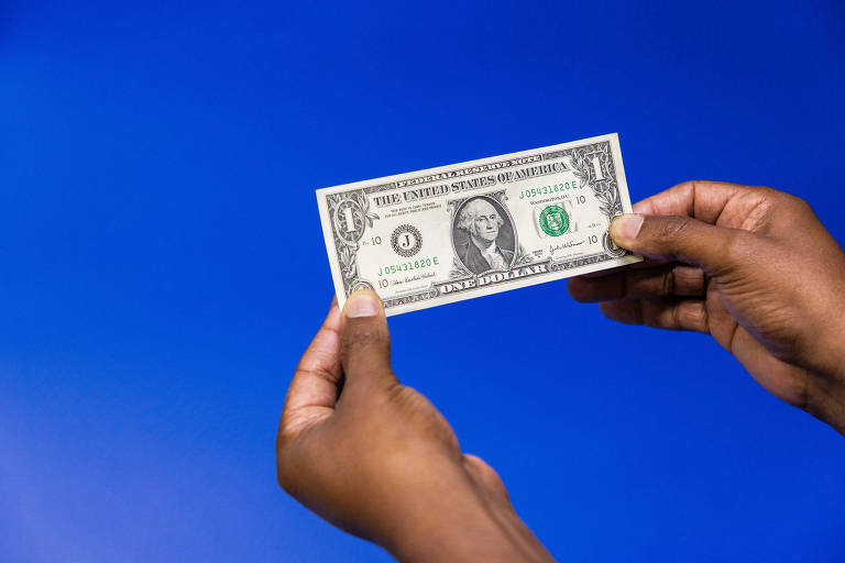 A imagem mostra uma nota de um dólar sendo segurada por duas mãos. O fundo é azul. A nota apresenta a figura de George Washington e o texto 039;THE UNITED STATES OF AMERICA039;.