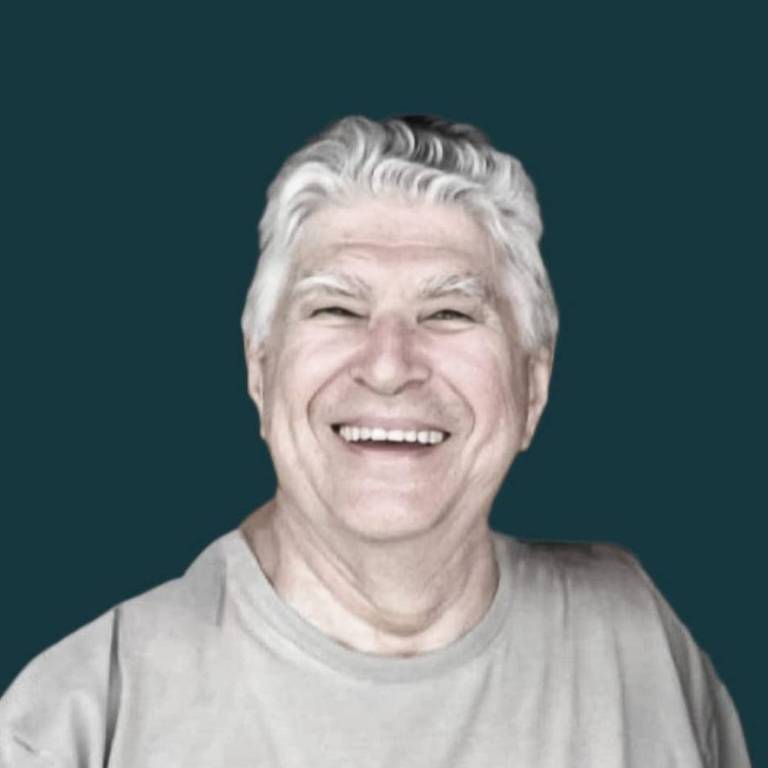 A imagem mostra um homem idoso com cabelo grisalho e um sorriso largo. Ele está usando uma camiseta clara e o fundo é de cor verde escura.