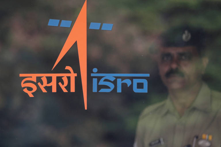 A imagem mostra o logotipo do ISRO (Organização de Pesquisa Espacial da Índia) em cores laranja e azul， com a palavra 039;ISRO039; escrita em inglês e em hindi. Ao fundo， há a silhueta de um guarda em uniforme.