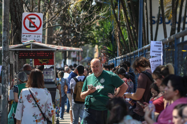 A imagem mostra uma fila de pessoas em uma calçada， com árvores ao fundo. À esquerda， há um sinal de proibição de estacionamento. Um homem de camiseta verde está em destaque， enquanto outras pessoas estão alinhadas， algumas olhando para seus celulares. Ao fundo， há uma estrutura que parece ser um quiosque ou uma cabine.