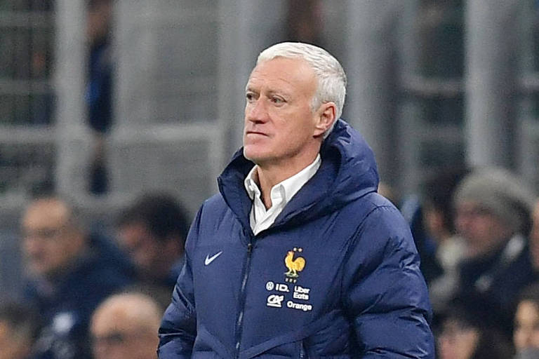 Um homem com cabelo grisalho e curto， vestindo um casaco azul escuro com o logotipo da seleção francesa de futebol， observa atentamente o jogo. Ele está em um ambiente de estádio， com torcedores ao fundo， e parece pensativo.
