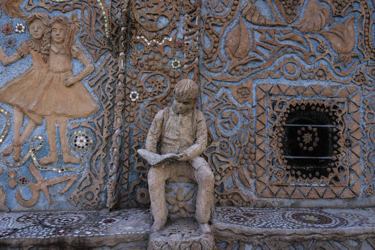 A imagem mostra uma escultura de um menino sentado， lendo um livro， em um mural decorativo. O fundo é composto por um padrão intrincado de texturas e cores， incluindo figuras de duas meninas em vestidos， além de elementos florais e geométricos. O menino está vestido com roupas simples e tem uma expressão concentrada enquanto lê. O mural é predominantemente em tons de azul e terracota， com detalhes em mosaico.