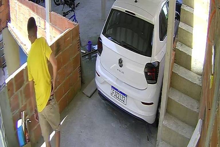 A imagem mostra uma garagem com um carro branco estacionado. À esquerda， uma pessoa vestindo uma camiseta amarela e shorts claros está de costas， olhando para uma parede de tijolos. Ao fundo， há uma bicicleta e outros objetos. A garagem tem uma escada à direita que leva a um andar superior.