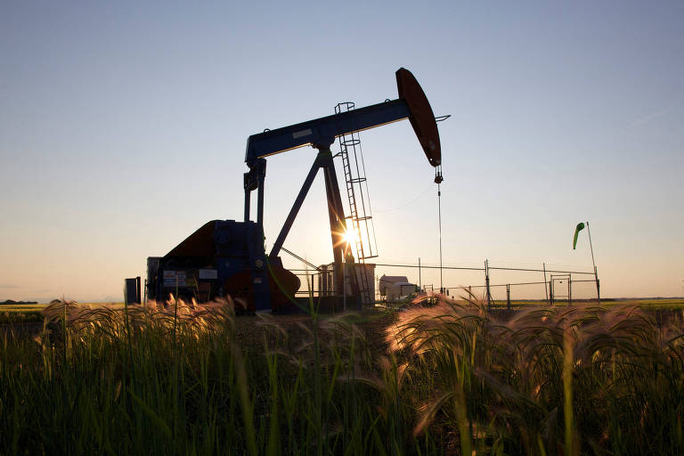 A imagem mostra uma bomba de petróleo em um campo， com o sol se pondo ao fundo. A estrutura da bomba é visível， e há uma cerca ao redor da área de extração. Na frente， há grama alta que se destaca contra o céu claro.