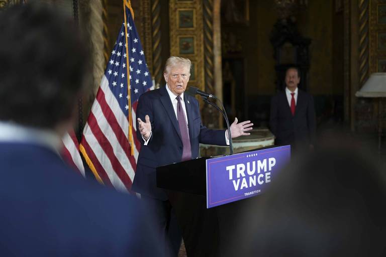 Na imagem， Donald Trump está em pé atrás de um púlpito com um cartaz que diz 039;TRUMP VANCE039;. Ele está gesticulando com as mãos enquanto fala. Ao fundo， há uma bandeira dos Estados Unidos e outra pessoa está parcialmente visível à direita. O ambiente parece ser um salão decorado.