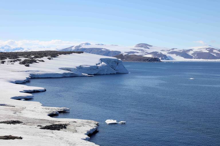 A imagem mostra uma paisagem com uma costa coberta de neve e gelo. O mar é de um tom azul profundo， refletindo o céu claro e ensolarado. Ao fundo， há montanhas cobertas de neve， criando um contraste com a água. Pequenos blocos de gelo flutuam na superfície do mar.