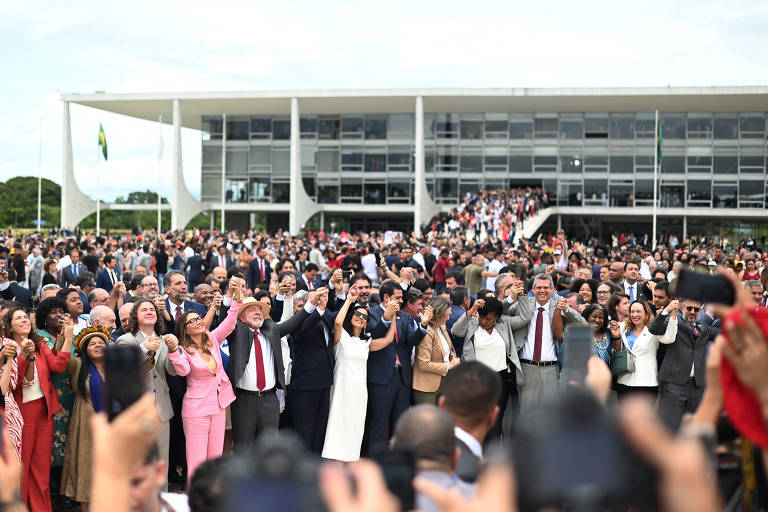 A imagem mostra uma grande multidão reunida em frente ao Palácio da Alvorada, com várias pessoas levantando os braços em celebração. O edifício moderno ao fundo é de arquitetura contemporânea, com janelas grandes e uma bandeira do Brasil visível. A multidão é diversa, com pessoas de diferentes idades e etnias, vestindo roupas formais e casuais. O céu está parcialmente nublado.