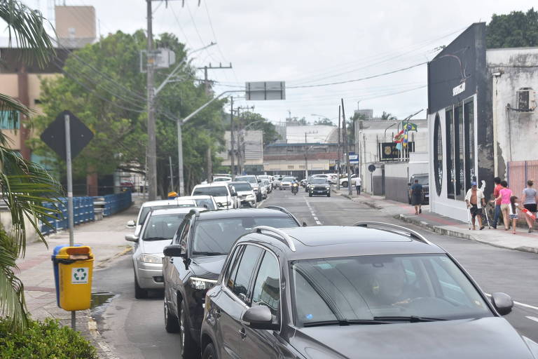 A imagem mostra uma rua urbana com tráfego intenso. Vários carros estão alinhados em uma fila， incluindo SUVs e sedãs. À esquerda， há uma lixeira amarela para reciclagem e postes de luz. Ao fundo， é possível ver um edifício e pessoas caminhando na calçada. O céu está nublado， sugerindo um dia com possibilidade de chuva.