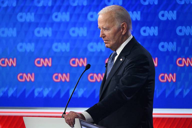 A imagem mostra um homem de cabelo grisalho， vestindo um terno escuro e uma gravata azul， em um cenário de debate. Ele está de perfil， olhando para baixo， com uma expressão séria. Ao fundo， há um painel com o logotipo da CNN em destaque， em um padrão azul e vermelho.