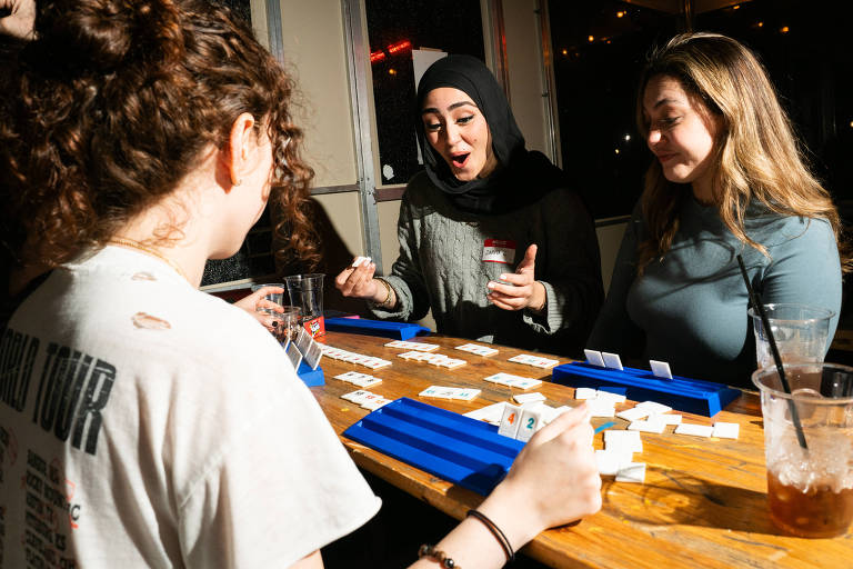 A imagem mostra um grupo de três pessoas jogando em uma mesa. Duas mulheres estão sentadas à direita， uma delas usando um hijab e a outra com cabelo solto. A mulher à esquerda， de costas， tem cabelo cacheado e usa uma camiseta branca. Elas estão interagindo animadamente enquanto jogam com peças dispostas em uma mesa de madeira. Copos de bebida estão visíveis na mesa.
