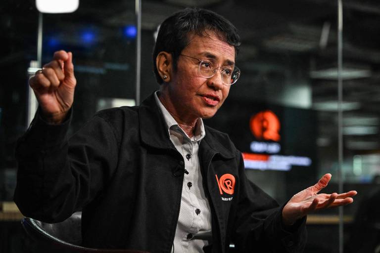 Imagem mostra Maria Ressa， uma mulher que está sentada em um estúdio， gesticulando com as mãos enquanto fala. Ela usa uma jaqueta preta com um logotipo visível. O fundo é desfocado， mas há luzes e elementos de um ambiente de estúdio de gravação.