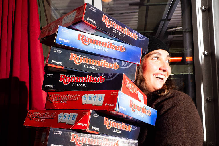 Uma pessoa está segurando uma pilha de caixas de jogos de tabuleiro, com destaque para a marca 'Rummikub'. A pessoa está sorrindo e parece estar em um ambiente iluminado, com uma parede vermelha ao fundo. As caixas estão empilhadas de forma instável, e a pessoa está usando um casaco escuro.