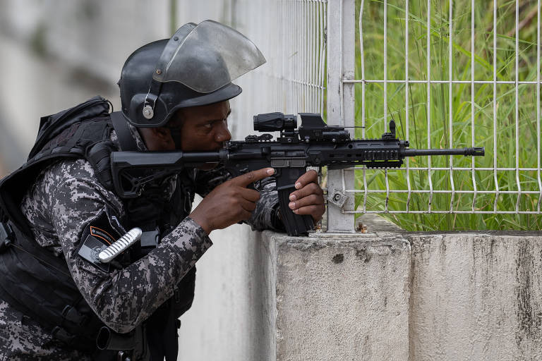 Um policial está posicionado atrás de uma barreira, mirando com um rifle. Ele usa um colete à prova de balas e um capacete, e está focado em um alvo além da cerca. O ambiente parece ser urbano, com uma grade ao fundo e vegetação visível.