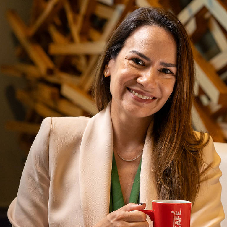 A imagem mostra uma mulher sorridente segurando uma caneca vermelha com a marca 039;NESCAFÉ039;. Ela está vestindo um blazer claro e uma blusa verde. O fundo apresenta uma estrutura de madeira emaranhada， criando um contraste com a sua roupa. A mulher tem cabelo longo e liso， e está em um ambiente bem iluminado.