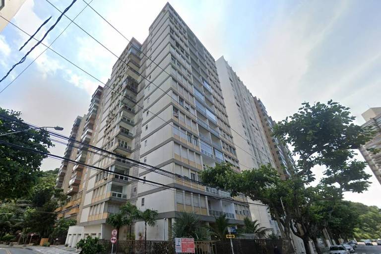 Imagem de um edifício residencial de vários andares， com uma fachada predominantemente clara e janelas grandes. O prédio está cercado por árvores e plantas， e há um calçamento em padrão na calçada. Fios elétricos estão visíveis no céu. Na parte inferior da imagem， há um sinal de trânsito e uma placa de aviso. O ambiente parece urbano e bem cuidado.