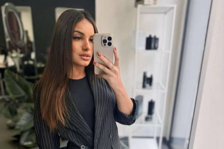 A imagem mostra uma jovem mulher tirando uma selfie em um salão de beleza. Ela está usando um blazer listrado preto e uma blusa preta， com meias-calças escuras. O fundo apresenta um espelho e prateleiras com produtos de beleza. A mulher tem cabelo longo e liso， e está segurando um celular na mão direita.