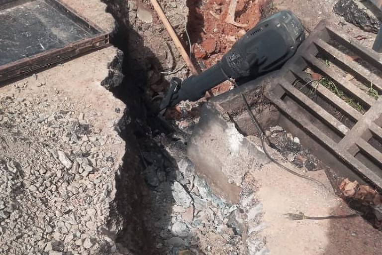 A imagem mostra um canteiro de obras com um buraco profundo no chão， cercado por pedras e materiais de construção. Há uma estrutura de metal ao fundo e uma escada de madeira ao lado. O solo está irregular e há uma tampa de bueiro visível.