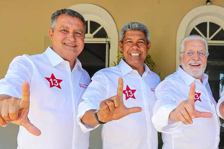 Rui， Jerônimo， Wagner estão posando para a foto， todos vestindo camisetas brancas com um símbolo vermelho em forma de estrela. Eles estão fazendo um gesto com as mãos， formando a letra 039;L039;. Ao fundo， há janelas com arcos e uma decoração simples.