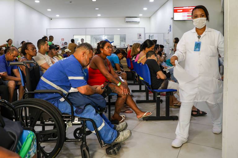 A imagem mostra uma sala de espera de um hospital com várias pessoas sentadas. À esquerda， um homem em uma cadeira de rodas está usando uma camisa azul e olhando para o celular. À sua frente， uma mulher está sentada em uma cadeira vermelha. No fundo， há várias outras pessoas sentadas e um profissional de saúde， vestido com um traje branco e máscara， caminhando em direção à câmera. A sala é bem iluminada e possui cadeiras azuis dispostas em filas.