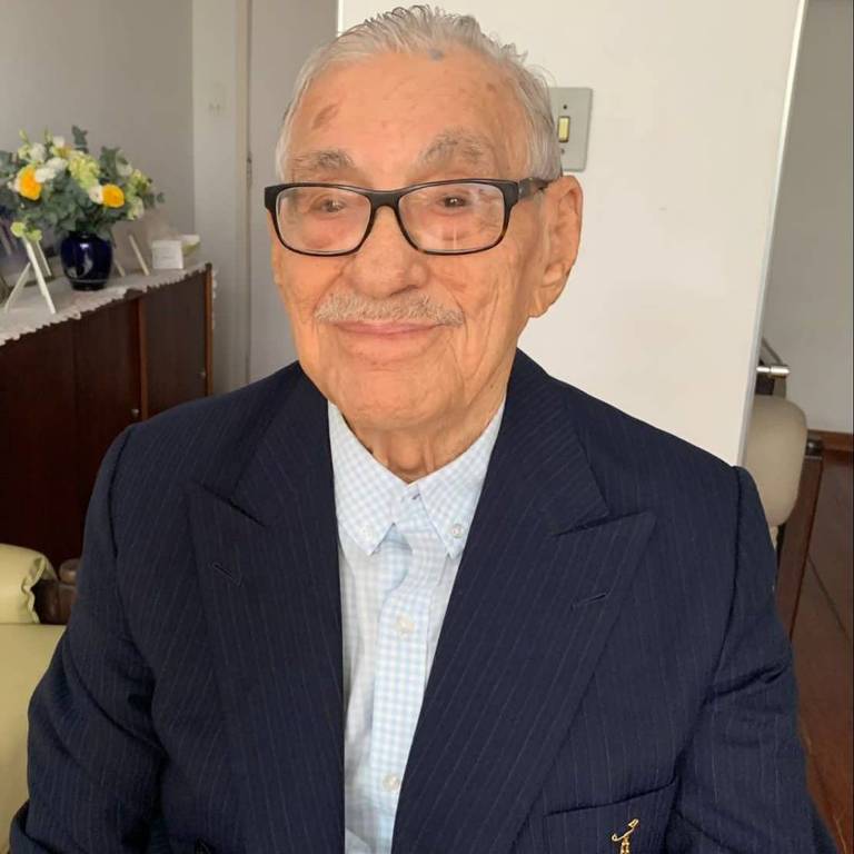 Um homem idoso está sentado, sorrindo para a câmera. Ele usa óculos e um terno escuro, com uma camisa clara por baixo. O fundo é um ambiente interno com móveis e flores visíveis.