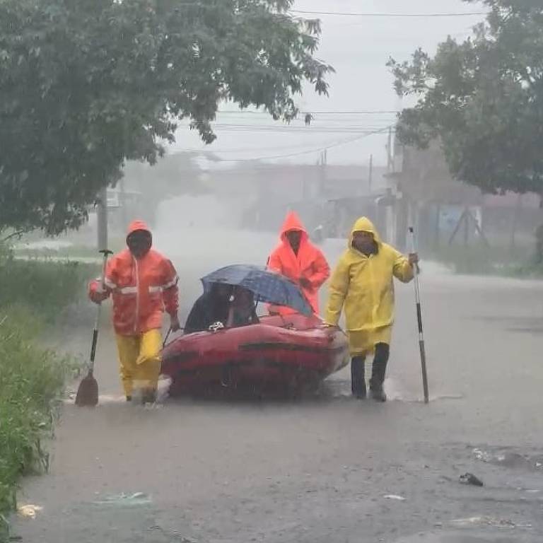 A imagem mostra três pessoas em uma área alagada， usando roupas de chuva. Duas delas estão puxando um barco vermelho， enquanto a terceira está ao lado， também vestindo um traje de proteção. O ambiente está chuvoso， com árvores ao fundo e uma estrada inundada.