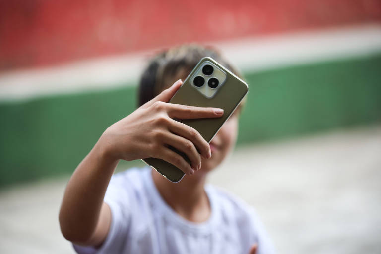 Uma pessoa está tirando uma selfie com um smartphone. A mão da pessoa segura o telefone em um ângulo que mostra seu rosto. O fundo apresenta uma parede com cores vermelha e verde.
