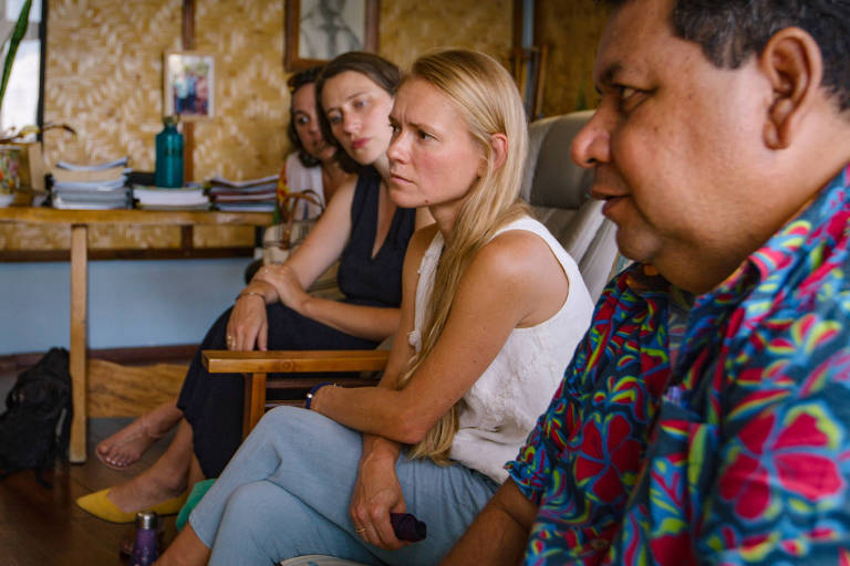 A imagem mostra um grupo de quatro pessoas sentadas em um ambiente interno. Duas mulheres estão à esquerda， uma com cabelo castanho e a outra com cabelo loiro longo. À direita， um homem com uma camisa colorida e uma mulher com cabelo castanho claro estão conversando. Ao fundo， há uma mesa com livros e uma garrafa de água. O ambiente tem paredes de madeira e uma decoração simples.