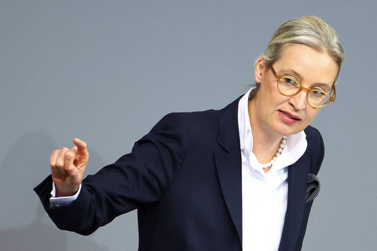 Uma mulher com cabelo loiro preso e óculos， vestindo um blazer escuro e uma camisa branca， está gesticulando enquanto fala. O fundo é de uma cor neutra， sugerindo um ambiente formal， possivelmente um discurso ou apresentação.