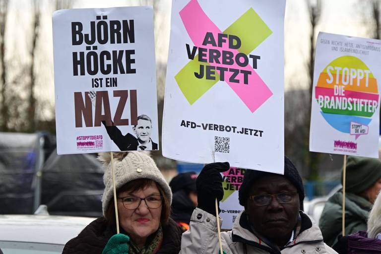 A imagem mostra duas pessoas segurando cartazes durante um protesto. A pessoa à esquerda usa um gorro e óculos， e o cartaz diz em alemão que 039;Björn Höcke é um nazista039;. A pessoa à direita tem pele escura e segura um cartaz também em alemão que pede 039;AfD proibida já039;. O ambiente parece ser ao ar livre， com árvores ao fundo.