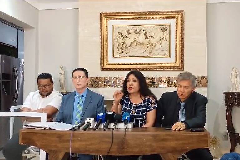 A imagem mostra uma coletiva de imprensa com quatro pessoas sentadas em uma mesa. À esquerda， um homem de camiseta branca. No centro， um homem de terno azul claro e gravata. Ao lado， uma mulher com blusa de bolinhas e cabelo solto， que parece estar falando. À direita， um homem de terno escuro. Ao fundo， uma parede com uma obra de arte e uma mesa decorativa.