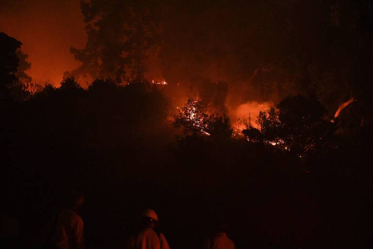 Imagem noturna mostra montanha sob chamas em Los Angeles， Califórnia. O céu escuro é tomado por um grande clarão laranja causado pelo fogo