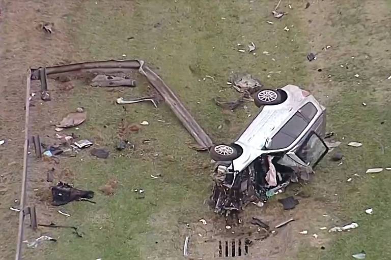 A imagem mostra um carro capotado em um campo， cercado por destroços e detritos. O veículo está de cabeça para baixo， com as rodas para cima. Há uma barreira de proteção danificada ao fundo e vários objetos espalhados pelo chão， incluindo partes do carro e outros detritos. Um cone de sinalização laranja está visível à esquerda da imagem.