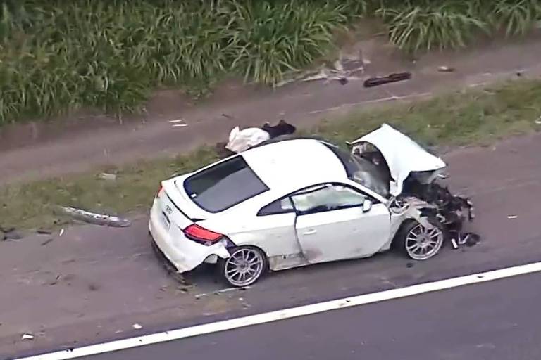 A imagem mostra um carro branco， modelo Audi， que sofreu um acidente. O veículo está danificado， com a parte traseira severamente comprometida e a porta do lado direito aberta. Há destroços espalhados ao redor do carro e um cone de sinalização na lateral da estrada. Ao fundo， uma pessoa está filmando a cena com uma câmera. A vegetação alta está visível ao lado da estrada.