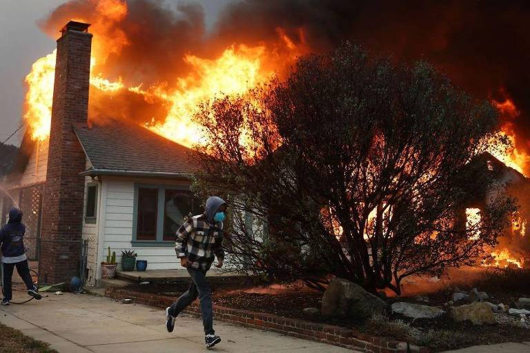 A imagem mostra uma casa em chamas， com grandes chamas e fumaça saindo do telhado. Duas pessoas estão visíveis: uma corre em direção à câmera， enquanto a outra está mais distante， aparentemente tentando apagar o fogo com uma mangueira. Há um arbusto próximo à casa， que também está sendo consumido pelo fogo.