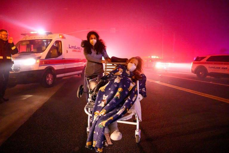 A imagem mostra uma cena de evacuação em uma situação de emergência. Uma mulher está sendo empurrada em uma cadeira de rodas por outra mulher， ambas usando máscaras. A mulher na cadeira de rodas está coberta com um cobertor. Ao fundo， há veículos de emergência， incluindo uma ambulância， com luzes piscando em meio a uma névoa densa e colorida. A atmosfera parece tensa e caótica.