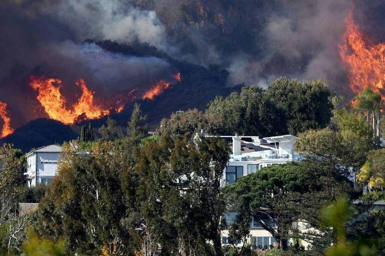 A imagem mostra um incêndio florestal em uma área montanhosa， com chamas intensas e fumaça densa subindo em direção ao céu. Na parte inferior， há casas cercadas por árvores， contrastando com o cenário devastador ao fundo. A vegetação está verde， mas as chamas estão consumindo a vegetação nas montanhas.