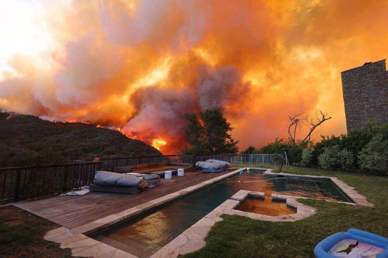 A imagem mostra uma área externa com uma piscina e um deck de madeira. No fundo， há um incêndio florestal， com grandes chamas e fumaça， iluminando o céu em tons de laranja e vermelho. À esquerda， há sofás cinzas e alguns objetos brancos. À direita， uma parede de pedra e vegetação ao redor.