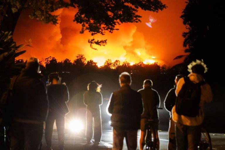 Um grupo de pessoas está de costas， observando um incêndio que ilumina o céu com uma luz laranja intensa. Algumas pessoas estão em pé， enquanto outras estão em uma bicicleta. A cena é noturna， com árvores ao fundo e uma fonte de luz vinda de um carro na frente.